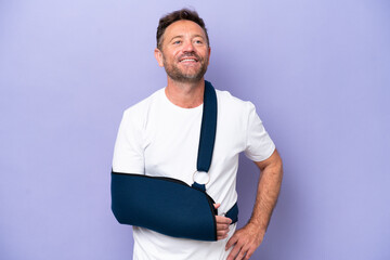 Middle age caucasian man with broken arm and wearing a sling isolated on purple background posing...