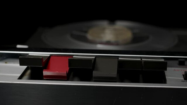 A vintage 1970s open-reel quarter-inch audio tape recorder spooling with record button depressed. Subdued lighting suggesting covert recording, wiretap, or espionage.