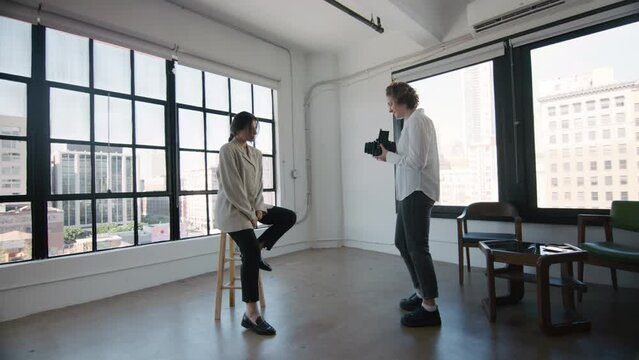 Young woman in casual white shirt and blue jeans, professional or hobby photographer using retro vintage film medium format camera, taking portrait picture, talking with model in photo studio in LA. 