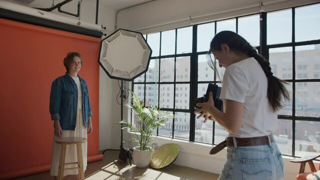 Young woman in casual white shirt, blue jeans, professional photographer using retro vintage film medium format camera, taking picture of model in denim shirt on terracotta background in photo studio