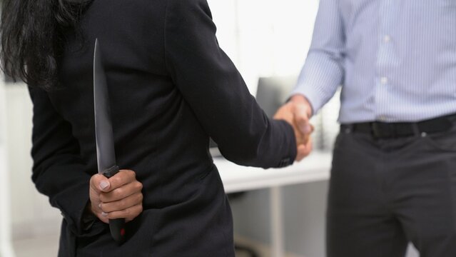 Back View Of Businesswoman Shaking Hands With Another Businessman While Holding A Knife Behind His Back. Concept Of Back Backstabbing In Business, Backstabbing Between Colleagues.