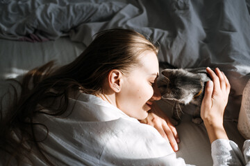 Candid portrait of Young woman is resting with kitten on the bed at home one sunny day. Girl play with outbred kitten.