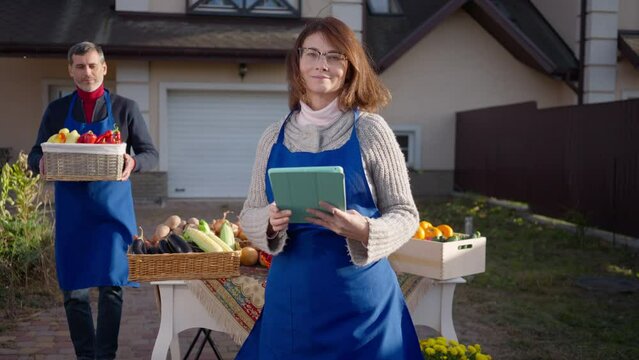 Portrait of beautiful confident woman surfing Internet on digital tablet looking at camera smiling as man bringing organic belly pepper in slow motion at background. Happy Caucasian farmers outdoors