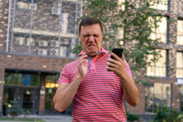 Dissatisfied millennial man in a pink T-shirt, chatting on the phone, looking at a gadget with an offended and disgusted expression, receiving a bad message.