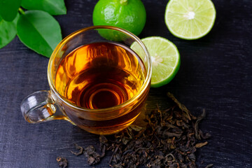 Hot tea in glass cup with fresh lime and green leaf on black wooden background.