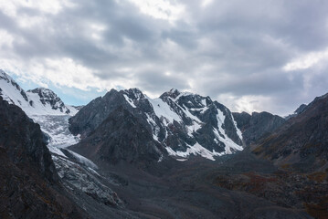 Fototapeta premium Atmospheric landscape with large snow mountain range with glacier and icefall in dramatic cloudy sky. Awesome high snowy mountains under rainy clouds. White snow on black rocks in overcast weather.