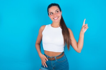 Smiling beautiful brunette woman wearing white tank top over blue background indicating finger empty space showing best low prices