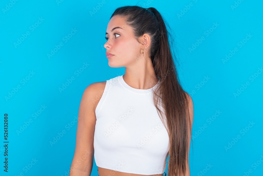 Wall mural Close up side profile photo beautiful brunette woman wearing white tank top over blue background not smiling attentive listen concentrated