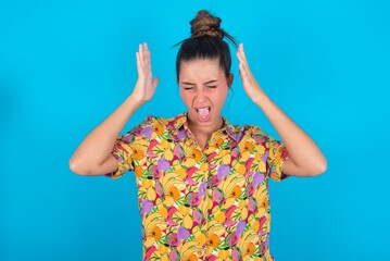 beautiful brunette woman wearing colourful shirt over blue background goes crazy as head goes...