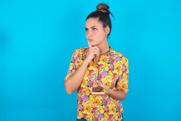 beautiful brunette woman wearing colourful shirt over blue background thinks deeply about...
