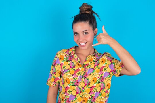 beautiful brunette woman wearing colourful shirt over blue background makes phone gesture, says call me back again, has glad expression.