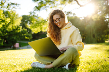 Beautiful woman  working online or studying at laptop sitting on grass at park. Business, blogging, freelance, education concept.