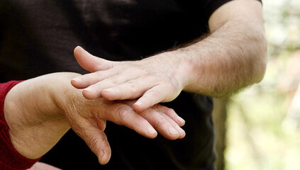 Close-up of tender gesture between two generations. Young man holding hands with a senior lady. Blurred background.