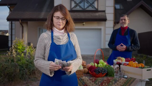 Portrait of Caucasian adult woman counting cash income posing standing on the left with blurred man selling local fruits and vegetables at background. Couple of farmers outdoors on autumn day