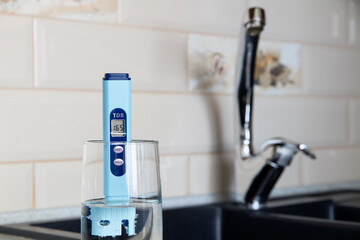 Electronic pH meter in a glass of water. In the background there is a tap for drinking water. The...