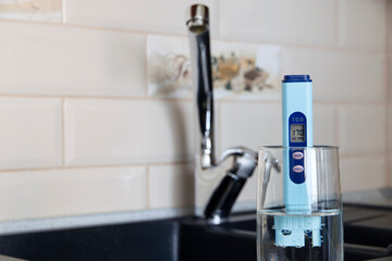  Electronic pH meter in a glass of water. In the background there is a tap for drinking water. The...