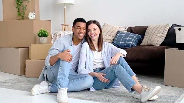 Happy multiracial family couple in love, hispanic guy and caucasian girl, sit on the floor in the living room of their new home, between cardboard boxes with household items, look at the camera, smile