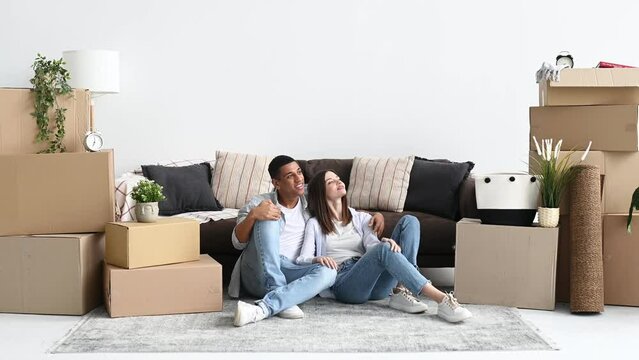 Happy spouses planning interior design of new home. Excited joyful mixed race couple in love, sitting on the floor in the living room in their new home, cardboard boxes with things are standing nearby