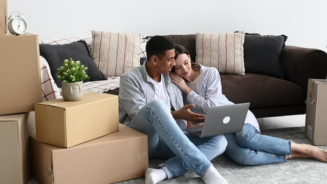 Happy married couple in love, man and woman of different nationalities, sit on the floor between boxes in their new housing, browse furniture in online stores, plan the interior of the apartment,smile