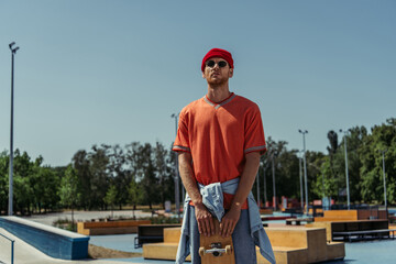 trendy man with skateboard standing in skate park in sunglasses and red beanie