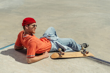 young man in orange t-shirt and beanie resting near skateboard in park