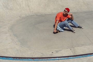 high angle view of man in red beanie and sunglasses sitting on skateboard in skate park