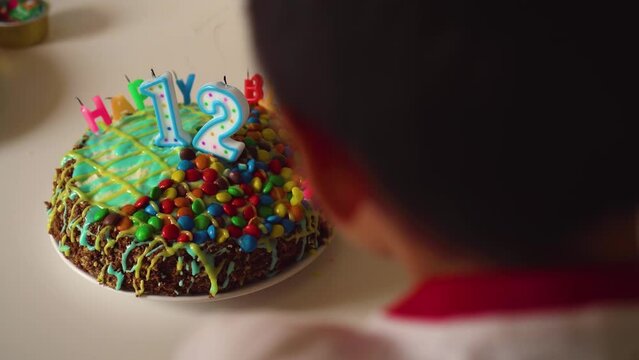 Cute boy blows out candles on sweet cake. Teenager celebrates twelve years birthday at home. Concept of happy childhood