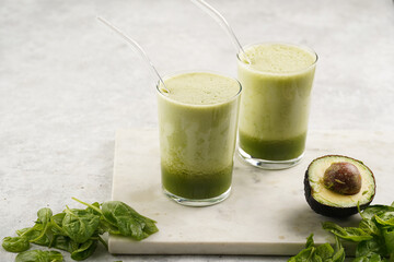 A glass of green healthy detox smoothie with spinach, matcha powder and a glass straw, fresh spinach and avocado on marble board on grey background
