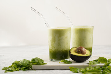 A glass of green healthy detox smoothie with spinach, matcha powder and a glass straw, fresh spinach and avocado on marble board on grey background