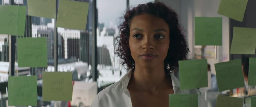 Young African American female employee looking at adhesive sticky notes on glass in the modern office, scrum agile methodology planning project. Shot through the glass
