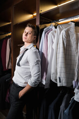 Elegant man looking at camera near shirts in cupboard in wardrobe
