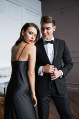Brunette woman in silk dress looking at camera near boyfriend in suit at home