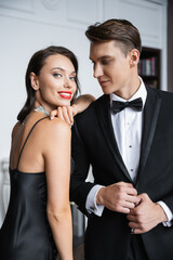 Smiling woman in dress looking at camera near elegant boyfriend at home