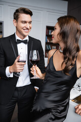 Smiling man in suit holding glass of wine near sensual girlfriend and piano at home