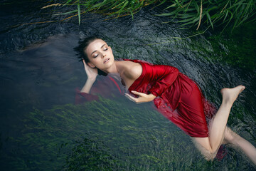 Art beautiful romantic portrait of a sexy young woman in a red dress lying in a river with green...