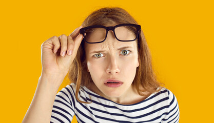Confused woman looks at you with suspicious, doubtful and incredulous expression on her face. Close up of skeptical and suspicious young woman looking out from under glasses on orange background.