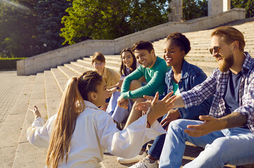 People hang out in summer. Group of cheerful friends talking, having fun and laughing sitting on stairs on street. Joyful multiracial young people listen to their female friend who tells funny stories