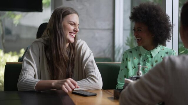 Three young diverse women laughing and smiling sitting at coffee shop table. Interaction between girlfriends in conversation. Real life laugh and smile