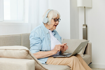 a happy modern old lady looks at the monitor holding a laptop on her lap talking through headphones smiling happily and gesticulating with her hands