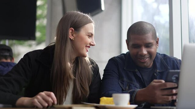 Young diverse couple seated at coffee shop looking at phone screen together. Happy people using smartphone sharing screen. Authentic real life laugh and smile