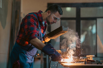 The welder performs welding task at his workplace in the factory, while the sparks 