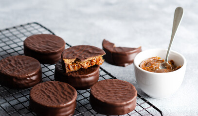 Dark chocolate alfajores on a dark rack and a bowl with dulce de leche, on grey background.  - obrazy, fototapety, plakaty