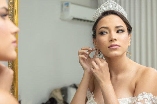 Jovem mulher vestida de noiva, de frente ao espelho, colocando seus brincos.