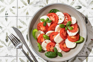 Italian caprese salad with sliced tomatoes, mozzarella cheese, basil, olive oil in green plate with cutlery on printed tile background. Top view