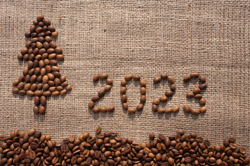 Merry Christmas and New Year greeting card. numbers 2023 and Christmas tree are made from coffee beans on fabric with burlap texture. View from above. New Year concept background