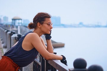 Portrait of a beautiful girl in sunglasses and cycling gloves at the railing of the embankment. The buildings and rotunda are blurred in the background. The concept of sport and healthy lifestyle.