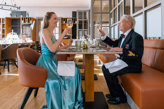 Eine junge, blonde Frau in einem hellblauen Abendkleid sitzt einem deutschen Marineunteroffizier in Gesellschaftsanzug gegenüber. Beide prosten sich mit einem Glas Weißwein zu. 