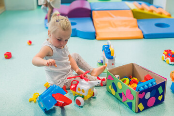 Toddler girl playing with different plastic toys such as building blocks, car toys. Work on...