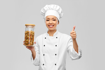 cooking, culinary and food concept - happy smiling female chef in toque holding jar with pasta and showing thumbs up gesture over grey background