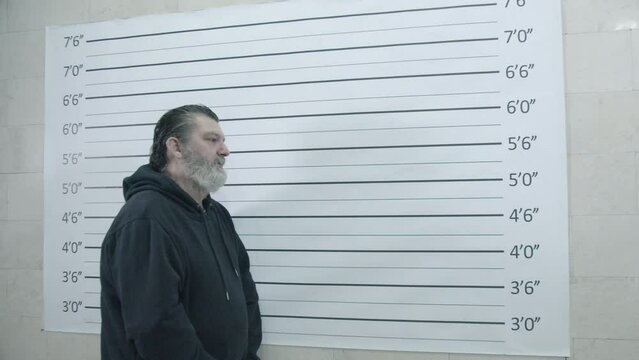 Portrait of the arrested caucasian mature man metric line wall during mug shot scene in a police station. Crane shot, slow-motion.